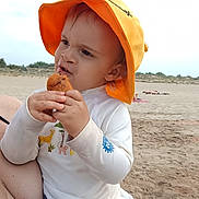 Izao a rejoint le concours — aidez-le/la à gagner de superbes lots ! toddler, child, orange_hat, snack, beach, sand, towel, white_shirt, outdoor, sunhat, food, messy_face, curious_expression, young_child, summer, vacation, nature, sky, person, sitting