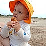 toddler, child, orange_hat, snack, beach, sand, towel, white_shirt, outdoor, sunhat, food, messy_face, curious_expression, young_child, summer, vacation, nature, sky, person, sitting