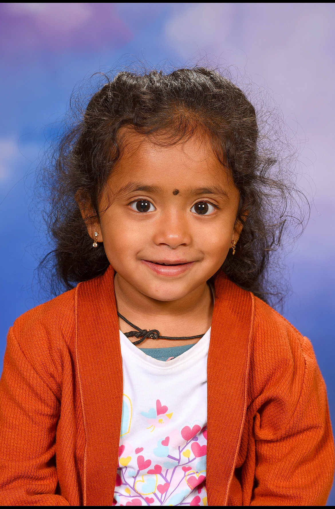 Nila participe au concours pour gagner de l'argent avec cette photo : child, girl, portrait, smile, curly_hair, orange_cardigan, earrings, necklace, colorful_shirt, hearts_pattern, studio_background, happy, cute, innocent, closeup, person, fashion, young, face, eyes