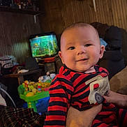 Easton is registered to the contest to win money with this photo: baby, infant, child, smiling, striped_pajamas, arm, hand, adult_hand, living_room, aquarium, fish_tank, toys, activity_center, blanket, plaid_blanket, couch, wood_paneling, shelf, radio, cozy_interior