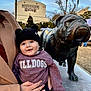 John is registered to the contest to win money with this photo: baby, child, person, statue, bulldog, bronze, outdoor, hat, sweater, smile, happy, hand, holding, building, lamp_post, tree, sky, maroon, black_hat, mississippi_state