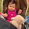 baby, child, stroller, teddy_bear, outdoor, grass, smiling, sitting, infant, pink_clothing, overalls, hair_bow, bottle, toy, portrait, playful, camping, wheel, fabric, family