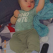 Eliel is registered to the contest to win money with this photo: baby, child, infant, bed, blanket, blue_blanket, clothing, dinosaur_print, green_pants, socks, remote_control, person, phone, hand, arm, face, lying_down, indoor, casual, cute