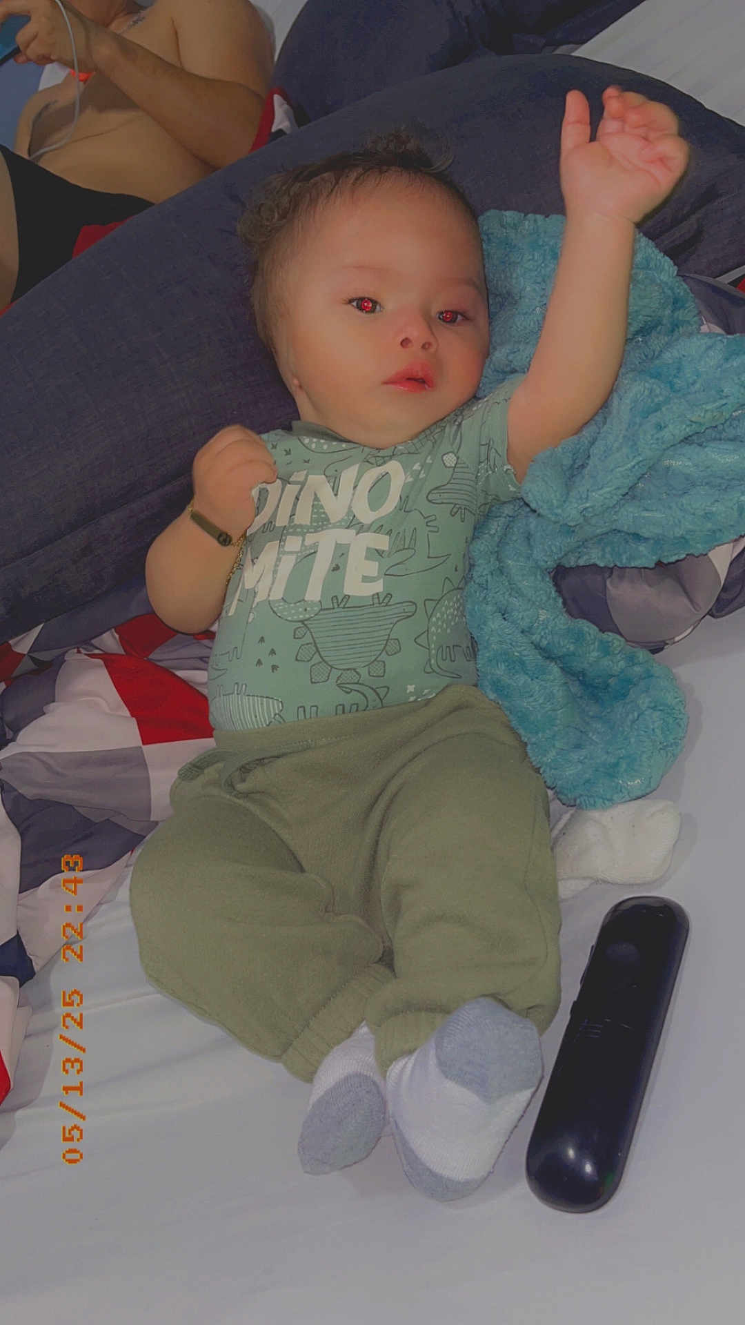 Eliel is registered to the contest to win money with this photo: baby, child, infant, bed, blanket, blue_blanket, clothing, dinosaur_print, green_pants, socks, remote_control, person, phone, hand, arm, face, lying_down, indoor, casual, cute