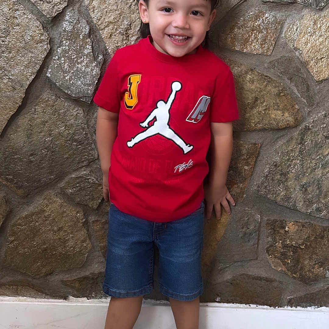 Angelrey is registered to the contest to win money with this photo: child, boy, smiling, standing, red_tshirt, denim_shorts, black_shoes, stone_wall, indoor, floor, happy, portrait, casual_clothing, short_hair, young, person, full_body, cute, fashion, playful