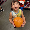child, ball, store, snacks, shelf, floor, drink, beverage, orange, tank_top, short_hair, curious, shopping, aisle, person, indoor, colorful, retail, food, young