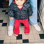 toddler, child, person, standing, jacket, red_pants, jeans, white_sneakers, floor, checkered_floor, indoor, hand, doorway, curly_hair, casual_clothing, cute, young_child, support, black_jacket, mickey_mouse_pattern