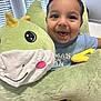 baby, child, smiling, plush_toy, dinosaur, blue_shirt, indoor, window_blinds, cute, happy, face, person, soft_toy, seated, infant, closeup, light, home, joy, playful