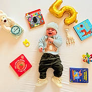 Aylan a rejoint le concours — aidez-le/la à gagner de superbes lots ! baby, smiling, teddy_bear, stuffed_animal, toy_giraffe, gold_balloon, books, board_books, teether, rattle, birthday_cake, knit_hat, sweater, corduroy_pants, white_sheet, sneakers, laying_down, bright_colors, happy, celebration