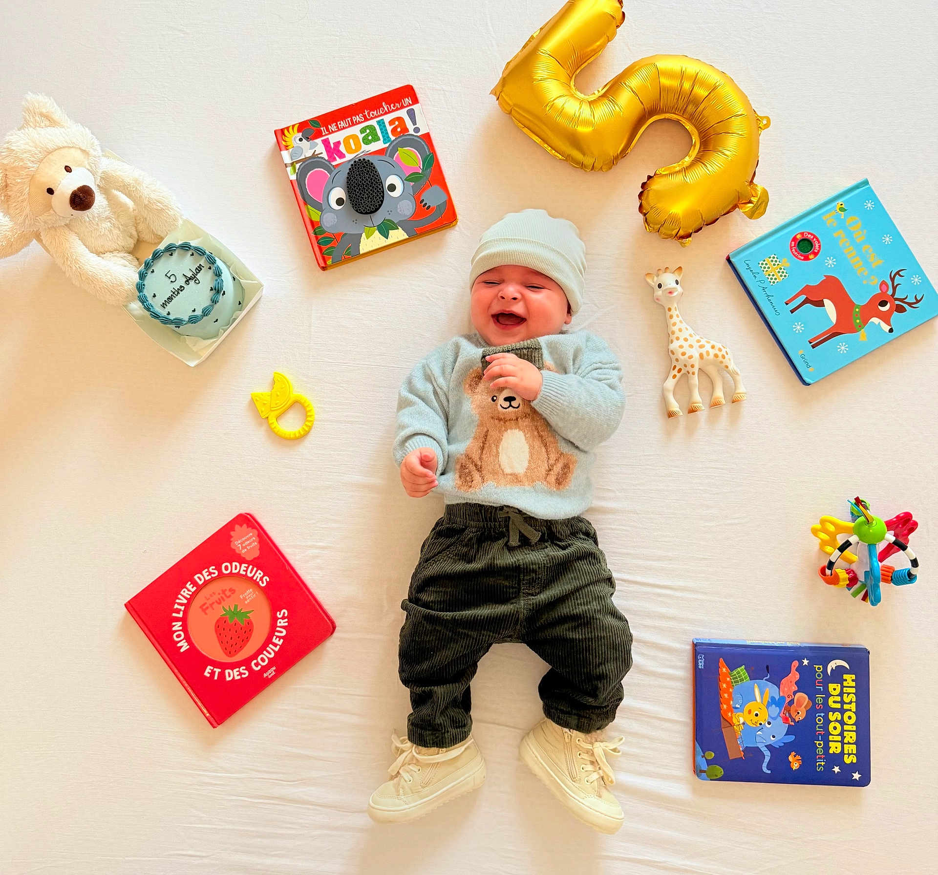 Aylan a rejoint le concours — aidez-le/la à gagner de superbes lots ! baby, smiling, teddy_bear, stuffed_animal, toy_giraffe, gold_balloon, books, board_books, teether, rattle, birthday_cake, knit_hat, sweater, corduroy_pants, white_sheet, sneakers, laying_down, bright_colors, happy, celebration