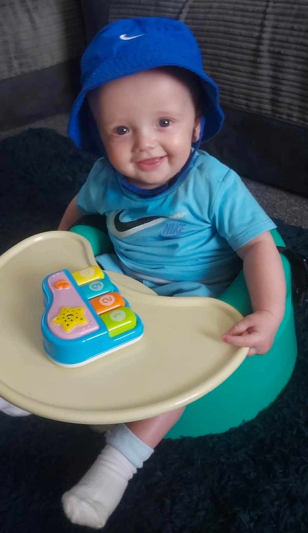 Tommy Rhys is registered to the contest to win money with this photo: baby, blue_hat, blue_shirt, nike_logo, baby_seat, toy_piano, smiling, indoor, child, playtime, sock, carpet, plastic_tray, happy, cute, face, infant, sitting, young_child, person