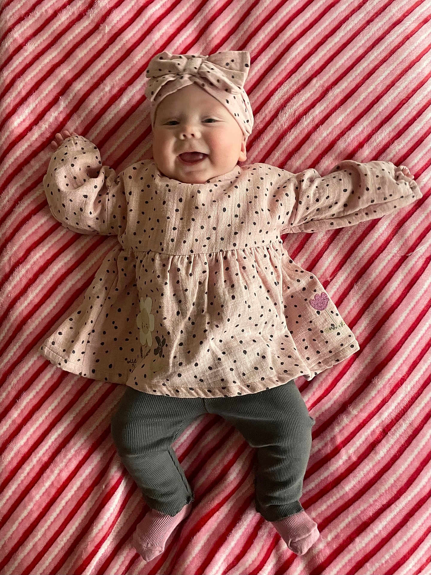 Bella-Rose is registered to the contest to win money with this photo: baby, infant, smiling, headband, pink_dress, polka_dots, striped_blanket, lying_down, happy, cute, child, toddler, cozy, soft_texture, patterned_clothing, indoors, feet, socks, portrait, playful