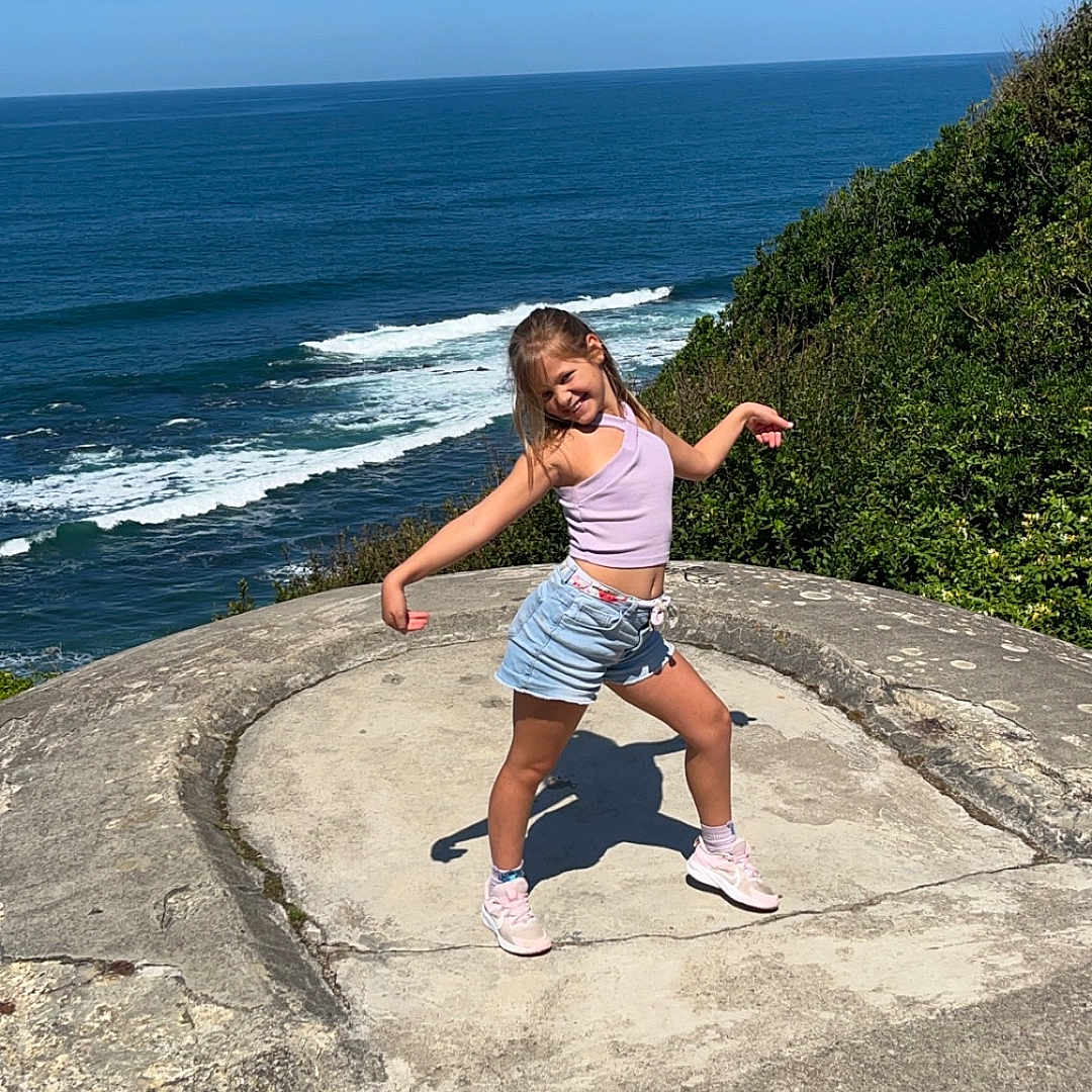 Lilou participe au concours pour gagner de l'argent avec cette photo : blue_sky, casual_clothing, child, concrete, girl, greenery, happy, ocean, outdoor, plants, playful, pose, sea, shorts, sky, sneakers, summer, sunlight, tank_top, waves