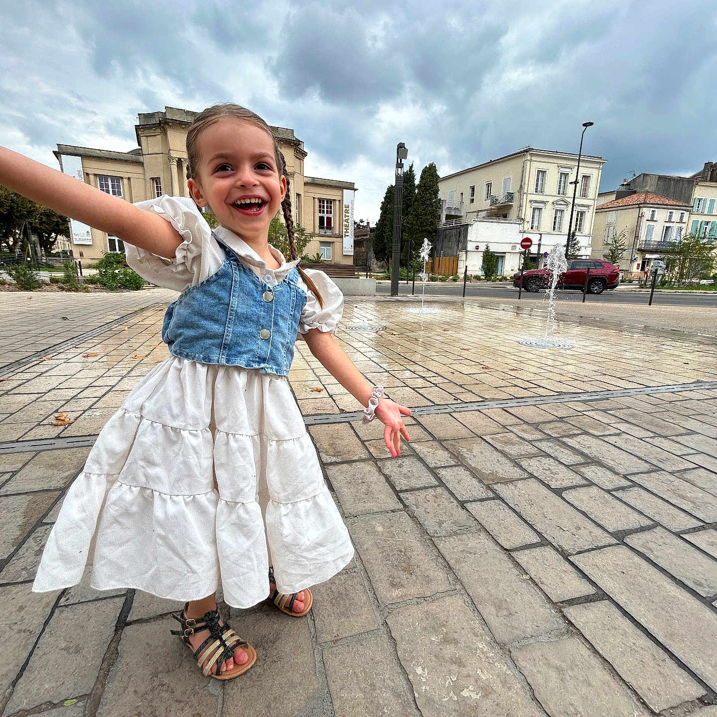 Syanna participe au concours pour gagner de l'argent avec cette photo : braids, buildings, child, cloudy_sky, daylight, dress, fountain, girl, happy, joyful, open_space, outdoor, pavement, person, playful, plaza, sandals, smiling, urban, water