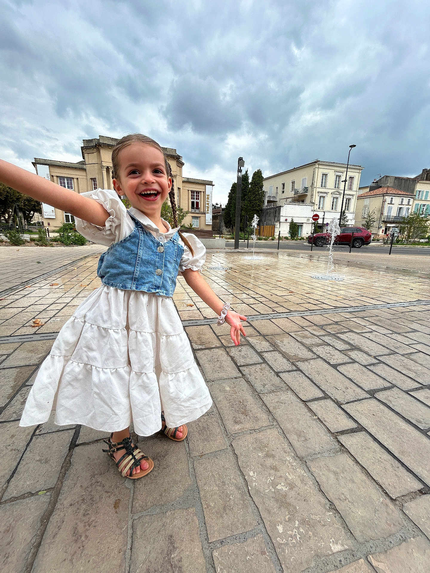 Syanna participe au concours pour gagner de l'argent avec cette photo : child, girl, smiling, braids, dress, sandals, outdoor, plaza, pavement, fountain, water, cloudy_sky, urban, buildings, happy, joyful, playful, person, daylight, open_space