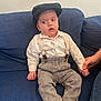 baby, infant, child, surprised_face, newsboy_cap, hat, suspenders, white_shirt, patterned_pants, socks, blue_couch, sofa, hand_holding, adult_hand, portrait, indoor, textured_wall, seated, expression, casual_clothing