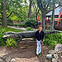 child, crocodile_statue, park, greenery, trees, rocks, pathway, wooden_stick, smiling, outdoor, casual_clothing, black_jacket, white_pants, shoes, daylight, playful, nature, fence, building, umbrella