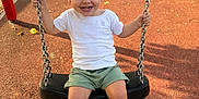 Kays participe au concours pour gagner de l'argent avec cette photo : child, toddler, swing, playground, outdoor, smiling, happy, white_shirt, green_shorts, white_shoes, metal_chains, sunlight, autumn_leaves, tree, fence, park, person, young_child, casual_clothing, daytime