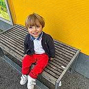 Achille a rejoint le concours — aidez-le/la à gagner de superbes lots ! child, blonde_hair, smiling, bench, yellow_wall, red_pants, white_shoes, outdoor, casual_clothing, happy, portrait, wooden_bench, asphalt, jacket, polo_shirt, sitting, daylight, playful, young_boy, urban