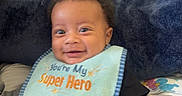 Eziah is registered to the contest to win money with this photo: baby, child, smiling, bib, clothing, black_clothing, indoor, pillow, text, happy, person, face, seated, adorable, infant, cute, young_child, black_hair, casual, home