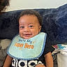 baby, child, smiling, bib, clothing, black_clothing, indoor, pillow, text, happy, person, face, seated, adorable, infant, cute, young_child, black_hair, casual, home