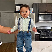 Maximiliano is registered to the contest to win money with this photo: child, toddler, baby, portrait, standing, kitchen, indoors, refrigerator, stove, microwave, cabinets, countertop, island, suspenders, shirt, jeans, sneakers, serious_expression, adult_hand, home