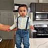 child, toddler, baby, portrait, standing, kitchen, indoors, refrigerator, stove, microwave, cabinets, countertop, island, suspenders, shirt, jeans, sneakers, serious_expression, adult_hand, home