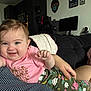 baby, smiling, pink_shirt, floral_pants, couch, blanket, indoor, person, child, happy, hand, face, living_room, gaming_chair, computer_monitor, wall_art, home, cozy, cute, portrait