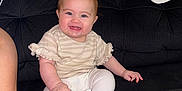 Rosalie is registered to the contest to win money with this photo: baby, child, smiling, sitting, toy, indoor, couch, black_couch, white_pants, beige_shirt, happy, cute, foot, hand, face, person, infant, home, blanket, cushion