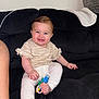 baby, child, smiling, sitting, toy, indoor, couch, black_couch, white_pants, beige_shirt, happy, cute, foot, hand, face, person, infant, home, blanket, cushion
