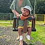 baby, child, swing, park, adult, green_dress, hat, bib, shoes, grass, trees, playground, fence, sunlight, footwear, person, outdoor, smile, holding, happy