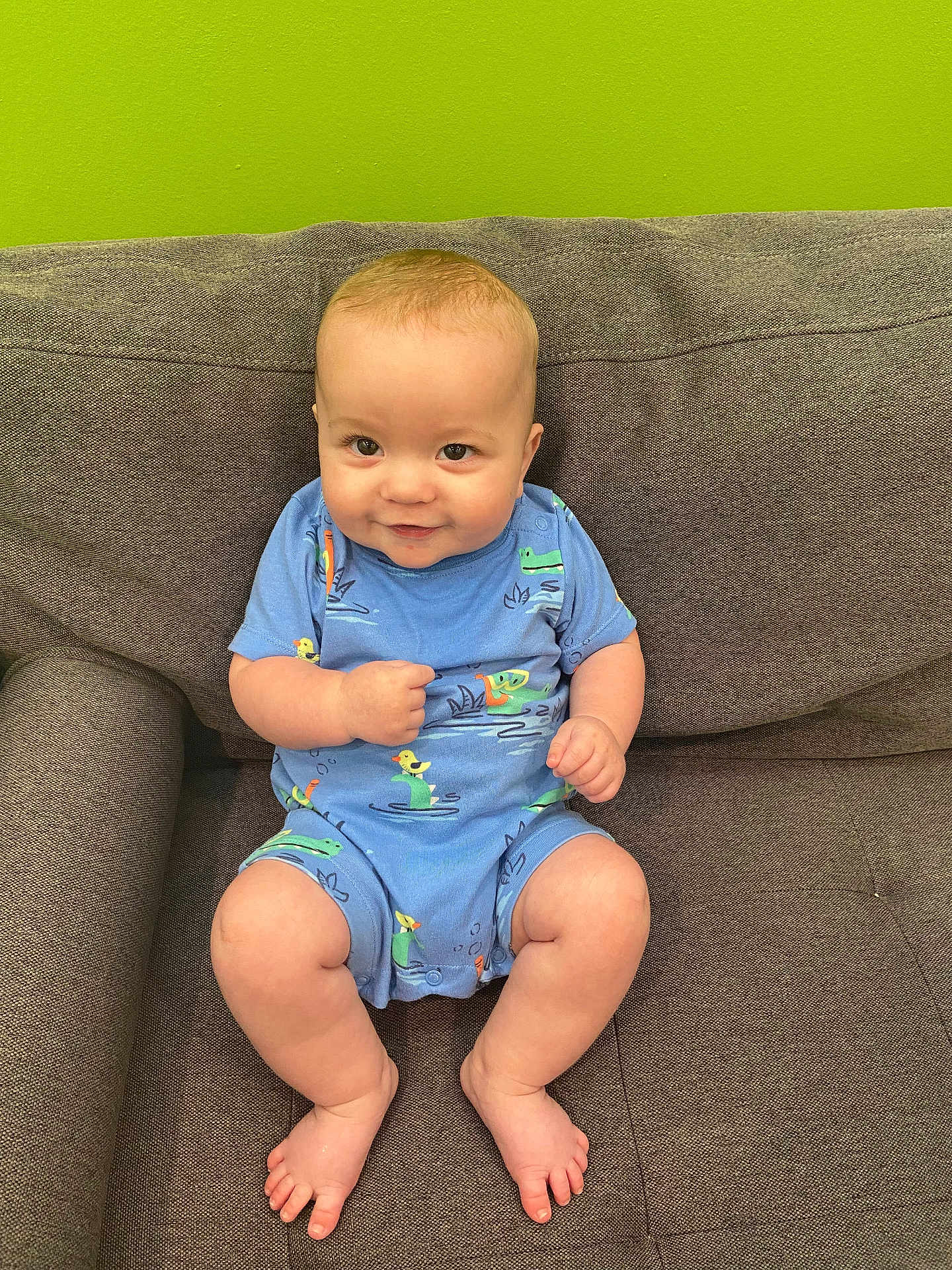 Roman is registered to the contest to win money with this photo: baby, child, smile, blue_onesie, couch, green_wall, sitting, barefoot, cute, infant, face, happy, plush, person, indoor, cozy, skin, legs, hand, expression