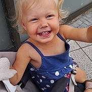 Noély participe au concours pour gagner de l'argent avec cette photo : child, toddler, smiling, happy, stroller, blonde_hair, pigtails, blue_dress, polka_dot, outdoor, seatbelt, footwear, pavement, wall, cute, baby, person, portrait, daylight, casual