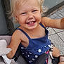 child, toddler, smiling, happy, stroller, blonde_hair, pigtails, blue_dress, polka_dot, outdoor, seatbelt, footwear, pavement, wall, cute, baby, person, portrait, daylight, casual