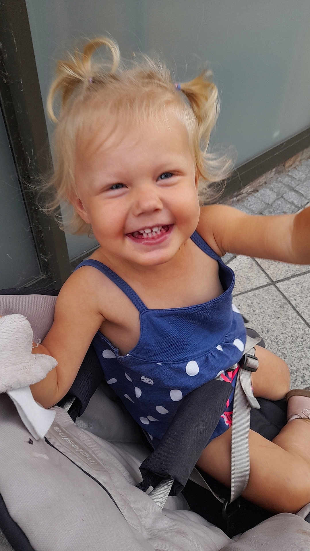 Noély participe au concours pour gagner de l'argent avec cette photo : child, toddler, smiling, happy, stroller, blonde_hair, pigtails, blue_dress, polka_dot, outdoor, seatbelt, footwear, pavement, wall, cute, baby, person, portrait, daylight, casual