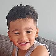 William joined the competition — help win amazing prizes! child, smiling, portrait, indoors, couch, gray_sofa, curly_hair, short_hair, tank_top, casual_clothing, happy, face, eyes, teeth, skin, sitting, home, close_up, photography, lifestyle