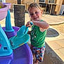 child, water_toy, outdoor, play, smile, green_shirt, colorful_shorts, water, purple_basin, blue_scoop, barefoot, sunlight, backyard, concrete, window, house, fun, happy, summer, toy