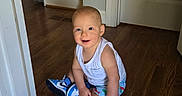 Sage is registered to the contest to win money with this photo: toddler, child, indoor, smiling, white_tank_top, shorts, blue_sneaker, barefoot, wooden_floor, doorway, home_interior, happy, sitting, cute, young_child, person, floor, light, casual_clothing, portrait