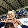 baby, child, person, hat, knit_hat, bear_ears, stadium, crowd, spectator, seating, adult, shoulders, clothing, outdoor, event, people, headwear, cute, warm_clothing, daytime