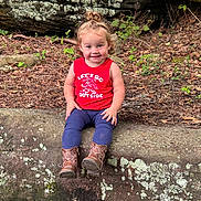 Delilah Mae is registered to the contest to win money with this photo: toddler, child, smiling, sitting, red_shirt, boots, rock, nature, outdoor, curly_hair, greenery, leaves, forest_floor, tank_top, happy, young_child, playful, casual_clothing, daylight, portrait