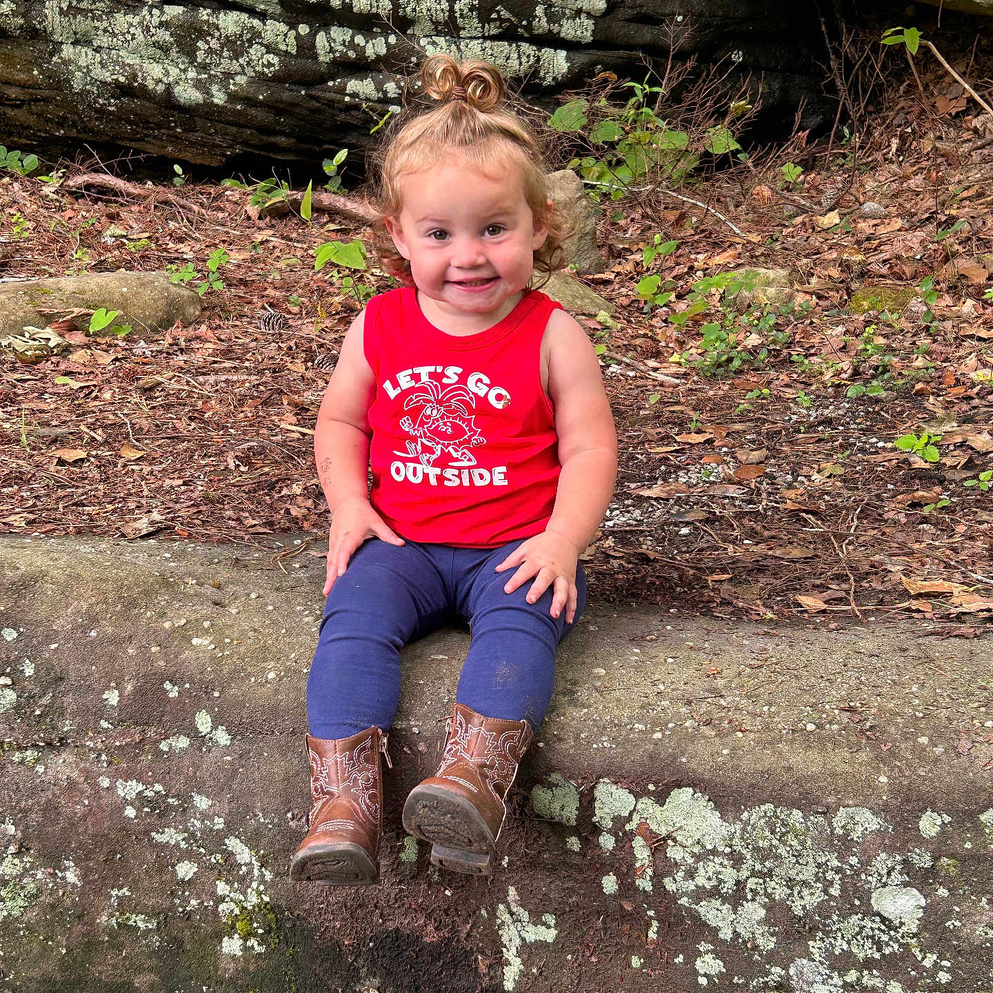 Delilah Mae is registered to the contest to win money with this photo: boots, casual_clothing, child, curly_hair, daylight, forest_floor, greenery, happy, leaves, nature, outdoor, playful, portrait, red_shirt, rock, sitting, smiling, tank_top, toddler, young_child