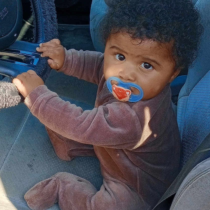 Khaïlly a rejoint le concours — aidez-le/la à gagner de superbes lots ! baby, brown_clothing, car_dashboard, car_door, car_interior, car_seat, child, curly_hair, cute, floor, footed_pajamas, hand, infant, looking_up, pacifier, seat_belt, seat_cover, steering_wheel, sunlight, window