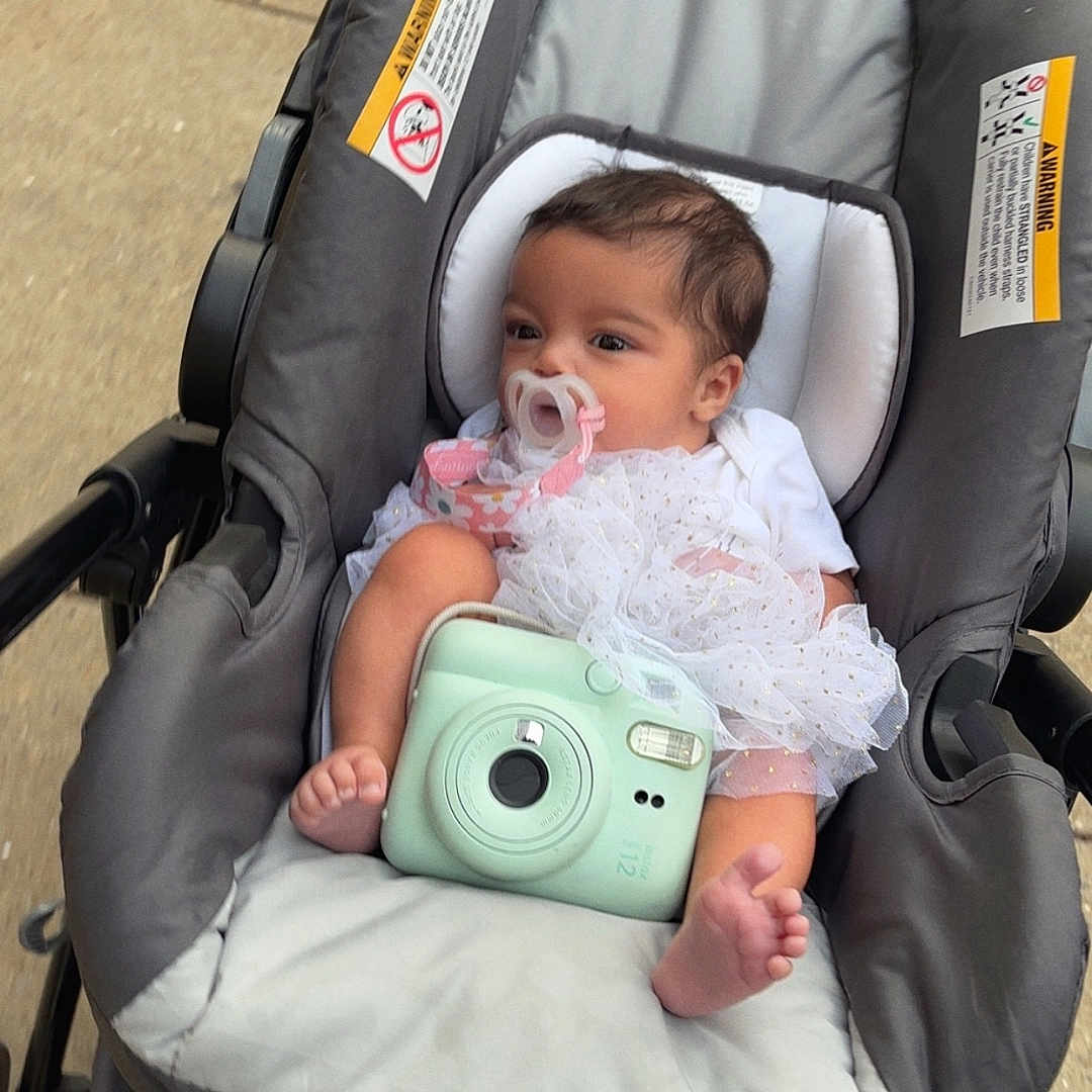 Solimar is registered to the contest to win money with this photo: baby, stroller, pacifier, camera, white_clothing, outdoor, child, infant, cute, seat, foot, head, toy, transport, person, baby_clothes, baby_accessory, baby_bottle_holder, infant_seat, baby_girl