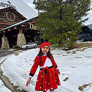 Noursan is registered to the contest to win money with this photo: child, girl, red_dress, knit_hat, polka_dot, snow, winter, pine_tree, log_cabin, outdoor_store, sidewalk, curb, parked_car, car, white_tights, red_shoes, smile, portrait, holiday_outfit, building