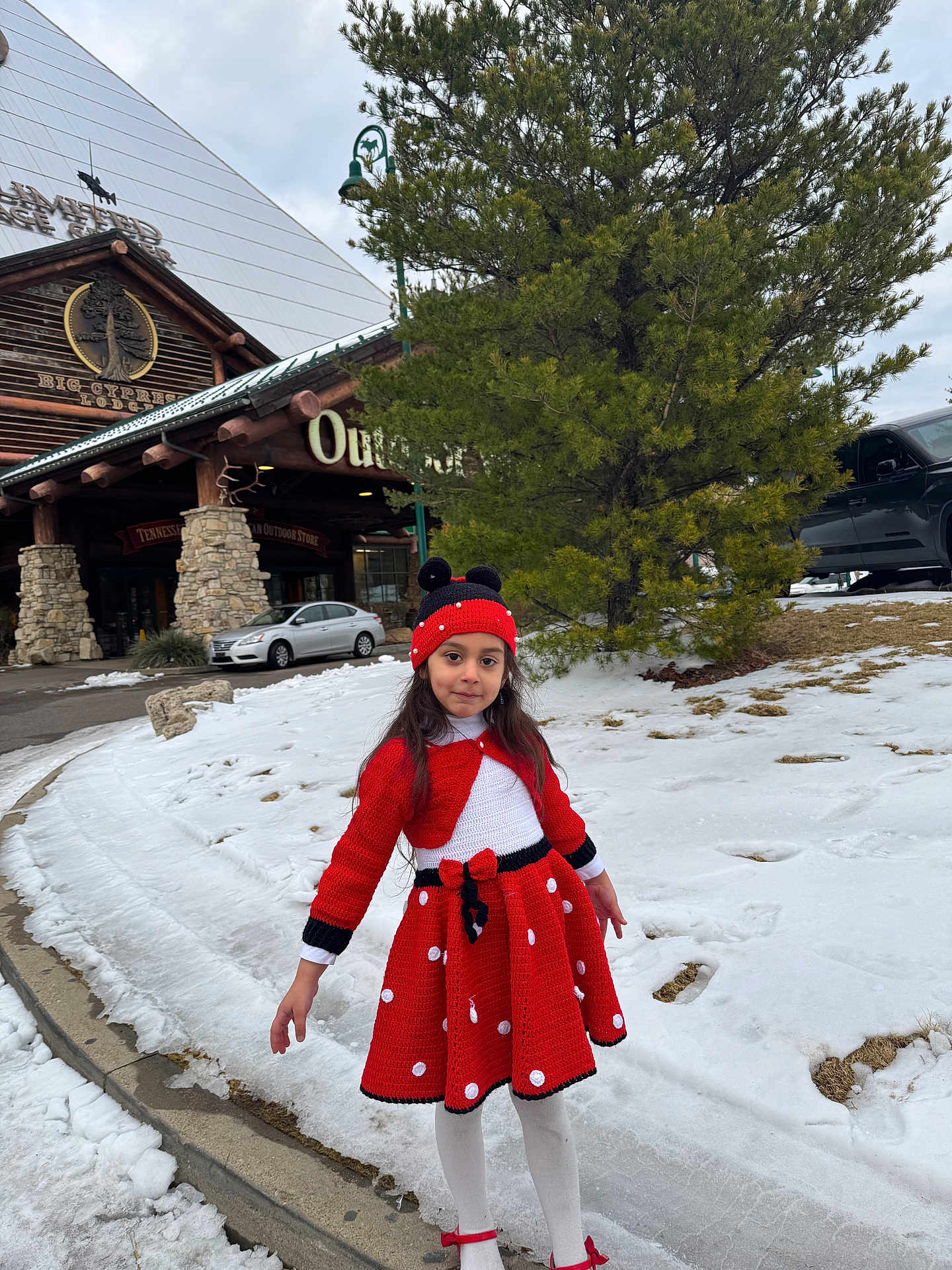 Noursan is registered to the contest to win money with this photo: child, girl, red_dress, knit_hat, polka_dot, snow, winter, pine_tree, log_cabin, outdoor_store, sidewalk, curb, parked_car, car, white_tights, red_shoes, smile, portrait, holiday_outfit, building