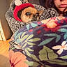 Poblo is registered to the contest to win money with this photo: dog, person, portrait, indoors, blanket, knitted_hat, winter_clothing, carrier, small_dog, wide_eyes, glasses, face, sitting, cozy, colorful_blanket, home, wood_floor, warm_lighting, pet, funny_expression