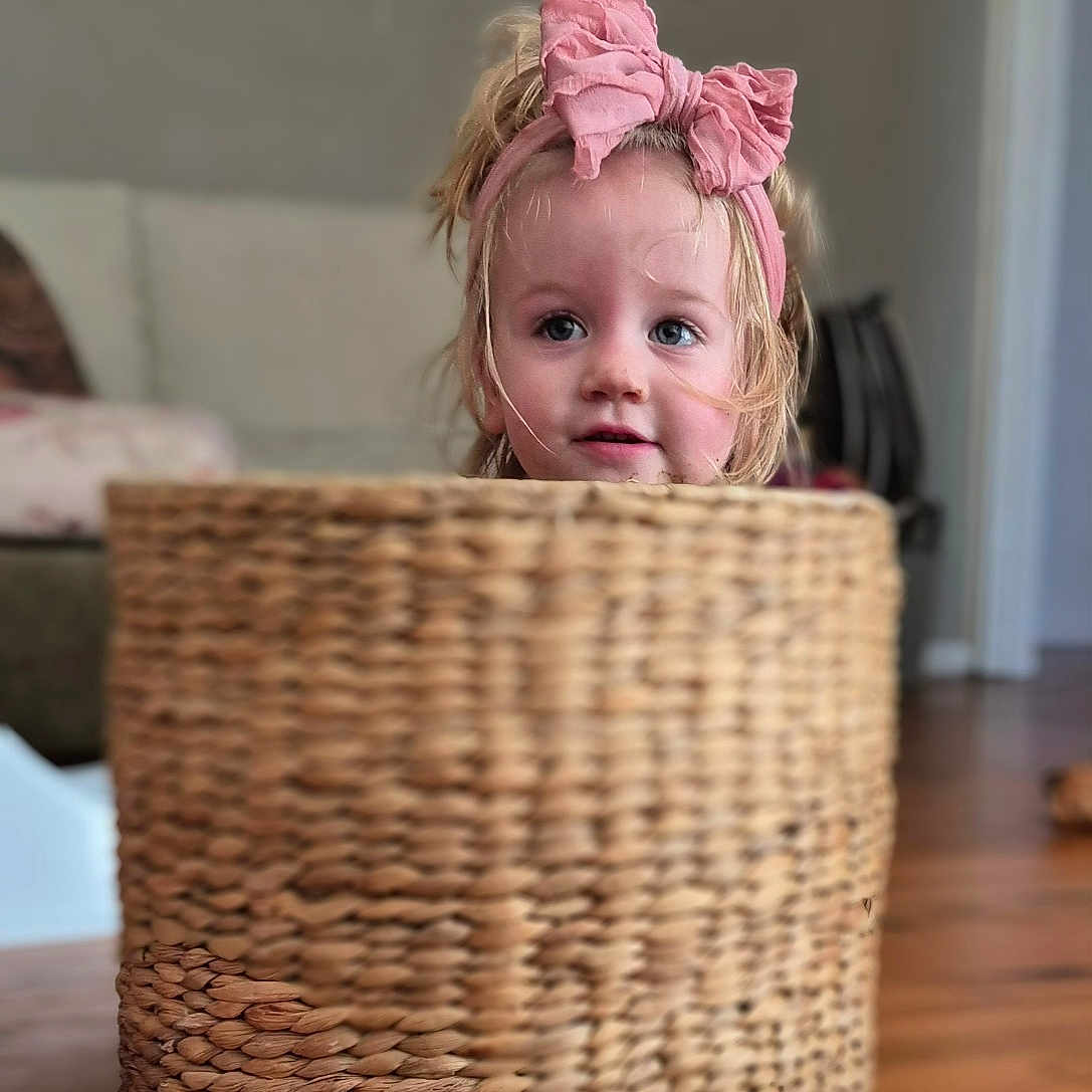 Ruby is registered to the contest to win money with this photo: accessories, baby, bag, basket, clothing, costume, couch, face, furniture, handbag, happy, hardwood, head, hiding, newborn, person, photography, portrait, smile, wood