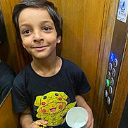 Ilyan a rejoint le concours — aidez-le/la à gagner de superbes lots ! child, boy, smiling, toy, cup, pikachu, tshirt, black_clothing, elevator, wooden_wall, buttons, indoor, portrait, young, person, casual_clothing, holding, standing, happy, cute