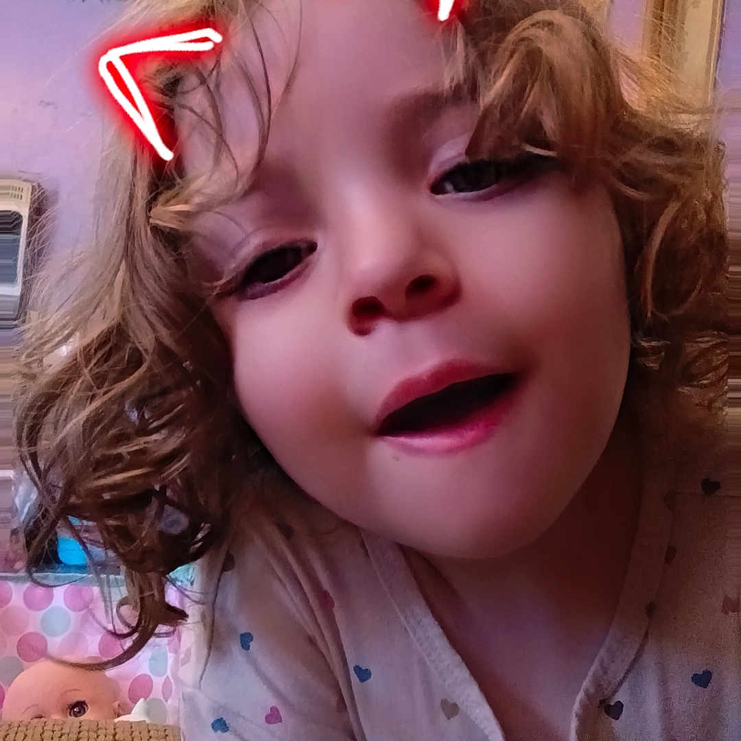 Ronni is registered to the contest to win money with this photo: child, curly_hair, face, indoor, neon_ears, playful, portrait, pajamas, person, cozy, home, doll, bedroom, curtains, window, smiling, cute, expression, closeup, casual