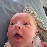Montie is registered to the contest to win money with this photo: baby, infant, close_up, face, expression, blanket, soft_texture, blue_background, newborn, curious, open_mouth, lying_down, skin, person, human, indoors, cute, head, small_hand, white_clothing